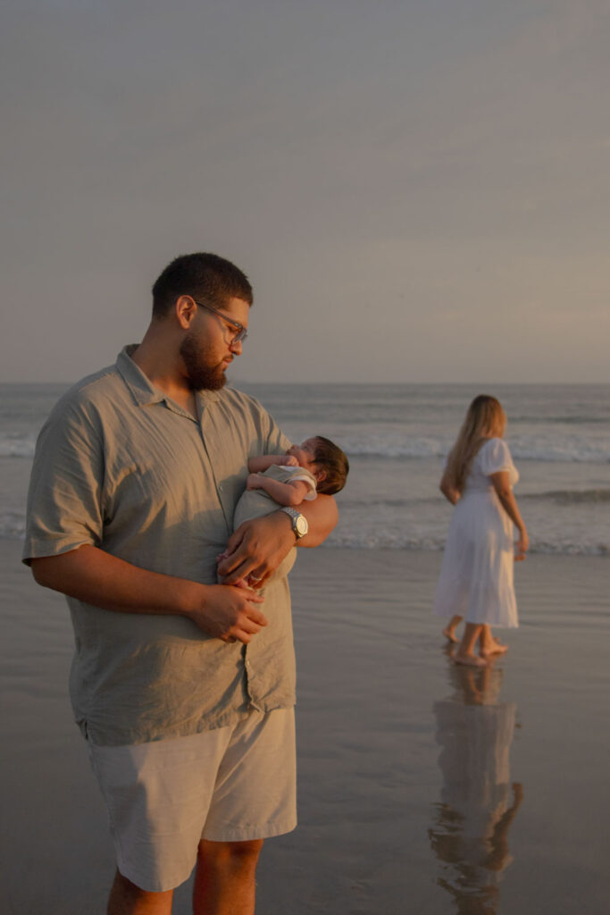 dad with glasses and light grey shirt holding his newborn baby on the beach with mom walking in the background